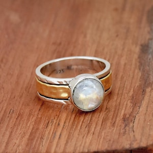 Op de afbeelding: Een zilveren ring met een gouden band en een ronde maansteen. De ring ligt op een houten oppervlak.