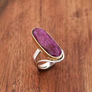 May include: A ring featuring a large, oval-shaped, deep pink gemstone set in a gold band. The band is surrounded by a silver band.