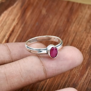 May include: A silver ring featuring a vivid oval ruby gemstone. The ring's band is slender and polished, with a simple design. The ruby is a deep red hue, set in a classic bezel setting. The ring is displayed on a wooden surface.