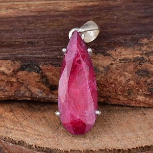 May include: A magenta-coloured, faceted, teardrop-shaped pendant. The gemstone is set in silver-coloured metal with a small loop for a chain. The pendant is displayed against a rustic, wooden background.