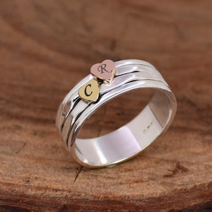 Puede incluir: Un anillo de plata con una banda texturizada y dos dijes en forma de corazón. Los dijes están grabados con las letras "C" y "R". Un dije es de oro y el otro es de oro rosa.