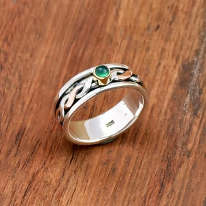 Może przedstawiać: Srebrny pierścionek z zielonym kamieniem szlachetnym i złotymi akcentami. Pierścionek ma pleciony wzór z miedzianymi detalami. Na obrączce widnieje oznaczenie "925". Pierścionek jest umieszczony na drewnianym tle.