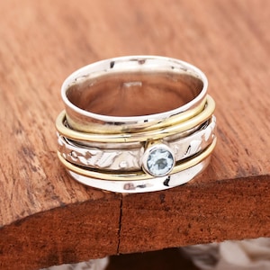 May include: A silver and gold spinning ring with a light blue gemstone. The ring has a textured design and is made of multiple bands.
