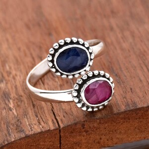 Puede incluir: Un anillo de plata con dos piedras preciosas ovaladas. La piedra más grande es un zafiro azul oscuro, y la piedra más pequeña es un rubí rosa. Ambas piedras están engastadas en un engaste de plata con un borde de cuentas.