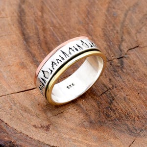 Peut inclure: Une bague en argent avec une bande rotative, ornée d'un motif de chaîne de montagnes. La bague a une bande dorée et une bande cuivrée. La bague est posée sur une surface en bois. La bague est marquée "925".