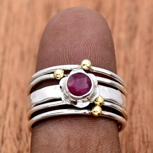 Puede incluir: Un anillo de plata con una piedra preciosa roja engastada en un diseño floral. El anillo tiene cuatro bandas y detalles dorados.