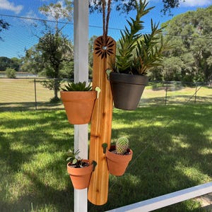 May include: A wooden hanging planter with four terracotta pots, holding various plants. The planter is made of cedar wood and has a sunflower design carved into it. The pots are filled with green plants, including succulents and a cactus.