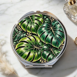 May include: A round, silver-toned compact mirror featuring a vibrant green monstera leaf pattern on its lid. The design displays detailed, overlapping tropical foliage with deep green and yellow-green hues, encased in a polished metallic frame.