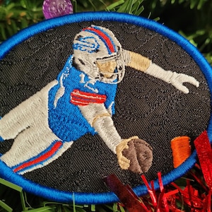 May include: A blue and white embroidered patch of a football player catching a ball. The patch is circular and has a blue border. The background is black.
