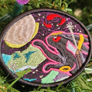 May include: A black embroidered patch with a colorful design of a creature with a red eye, a moon, and stars. The creature is surrounded by pink, green, and blue swirls.