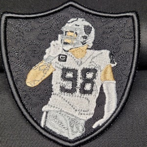 May include: Black and white embroidered patch of a football player wearing a helmet and jersey with the number 98. The patch is shaped like a shield.