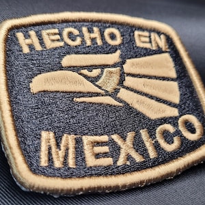 May include: Embroidered patch with the words "HECHO EN MEXICO" and an eagle design. The patch is square with a gold border and lettering, and the background is black. The eagle is tan and gold.