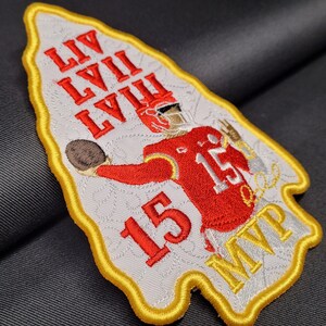 May include: A white and gold embroidered patch with a football player throwing a football. The patch features the text "LIV LVIII LVIII" and "15 MVP".