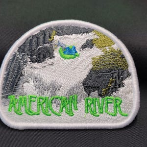 May include: White embroidered patch with a green outline. The patch features a stylized image of a river with a blue and green boat. The text "AMERICAN RIVER" is embroidered in green below the image.