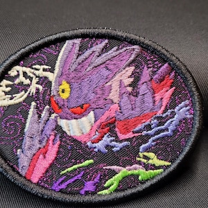 May include: Embroidered patch featuring a purple cartoon character with a wide grin, yellow eyes, and red accents. The patch has a black border and a dark background with colorful details. The design is oval-shaped.