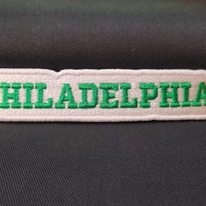 May include: White embroidered patch with the word "PHILADELPHIA" in green lettering.