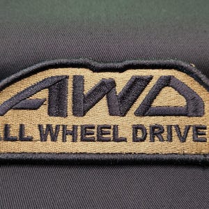 May include: A brown and black embroidered patch with the text "AWD ALL WHEEL DRIVE" in black.