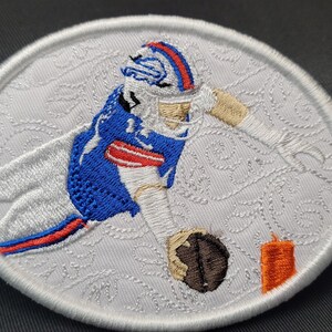 May include: White embroidered patch featuring a football player in a blue and white uniform, catching a football. The player is wearing a helmet and has a determined expression. The patch is surrounded by a white border.