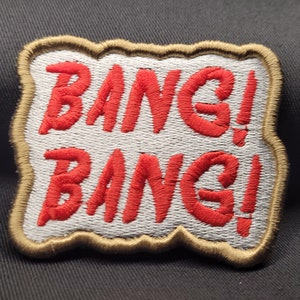 May include: A white embroidered patch with a red "Bang! Bang!" text in a comic book style font. The patch has a brown border.