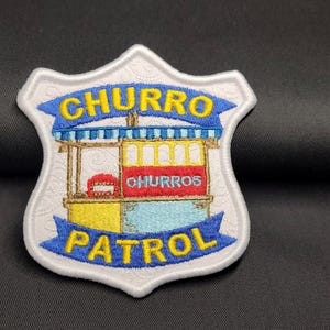 May include: Embroidered patch in the shape of a shield with the words "CHURRO PATROL" in yellow and blue. The patch features a churro stand with the word "CHURROS" in red. The patch is white with a black background.