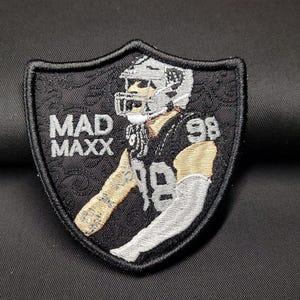 May include: Embroidered patch featuring a football player in a black and silver uniform, with the text "MAD MAXX" and the number "98". The patch has a shield shape with a decorative border.