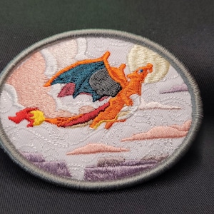 May include: Embroidered patch featuring a red and orange dragon with black wings flying in front of a white moon and pink clouds.