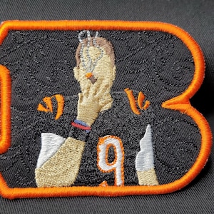 May include: An embroidered patch featuring a cartoon of a person wearing a black jersey with the number "9" on the front. The person is covering their face with their hand. The patch has an orange border.