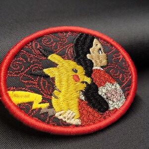 May include: Embroidered circular patch featuring Pikachu and a character in a red and black kimono. The patch has a red border and a black background with a red floral pattern. Pikachu is yellow with a red cheek and a lightning bolt tail.