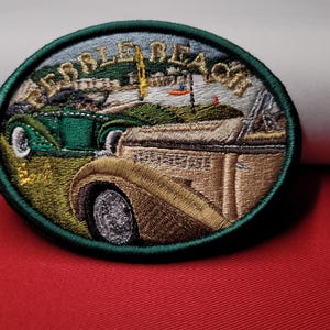 May include: Embroidered patch with the words "PEBBLE BEACH" in gold lettering. The patch features two vintage cars, one green and one tan, set against a coastal landscape with a golf course and ocean.