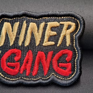 May include: Embroidered patch with the words "NINER GANG" in gold and red thread on a black background. The patch has a black border with gold stitching and is set against a dark gray fabric.