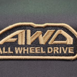 May include: Embroidered patch with the text "AWA ALL WHEEL DRIVE" in tan and black. The patch has a black background with tan stitching around the edges and the text. The patch is rectangular with rounded corners.