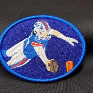 May include: Embroidered patch featuring a football player in a blue and white uniform diving towards the end zone. The patch is circular with a blue background and a blue border. The player is holding a brown football.