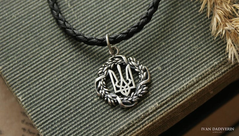 May include: A silver pendant necklace with a wheat wreath design surrounding the Ukrainian trident symbol. The necklace is on a black braided cord.