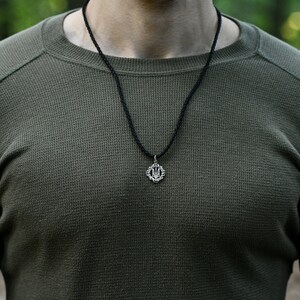 May include: A close-up of a person wearing a green long-sleeved shirt with a black cord necklace and a silver pendant featuring the Ukrainian trident symbol.
