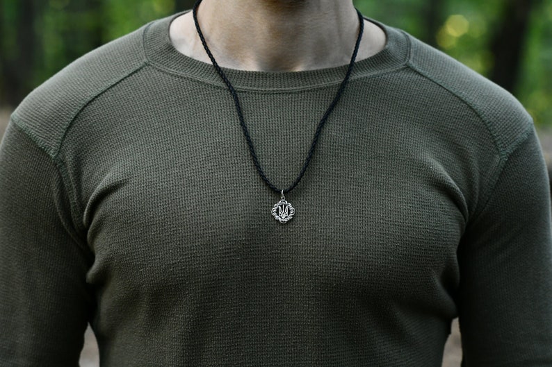 May include: A close-up of a person wearing a green long-sleeved shirt with a black cord necklace and a silver pendant featuring the Ukrainian trident symbol.