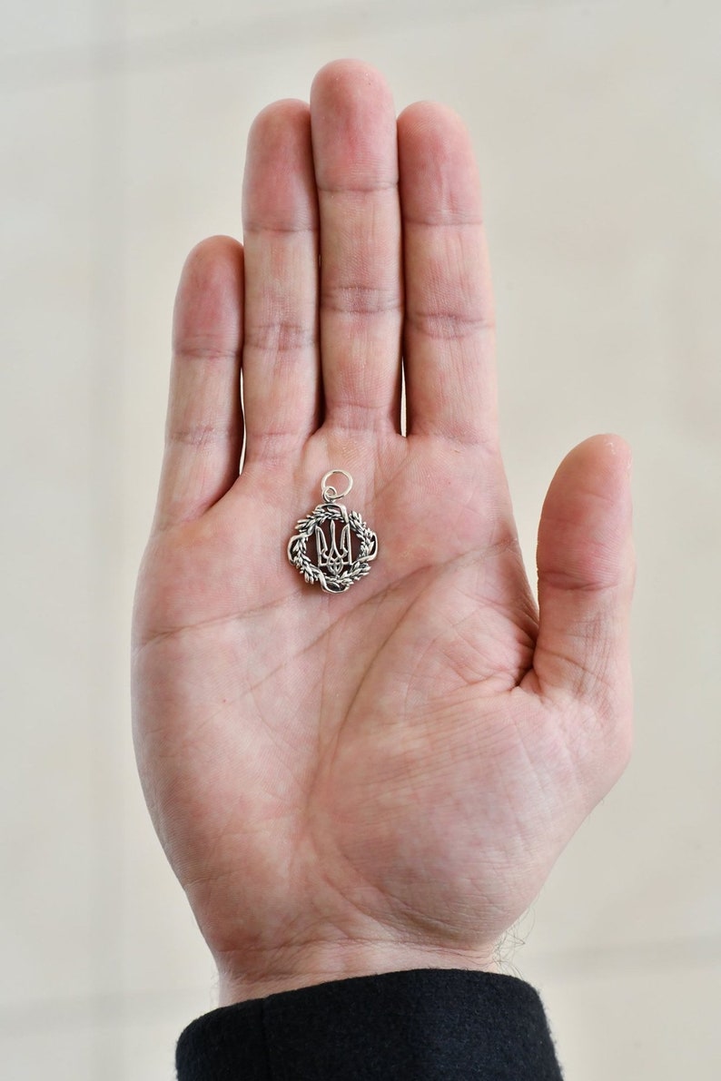 May include: A silver pendant featuring the Ukrainian trident symbol, surrounded by a wreath of leaves.