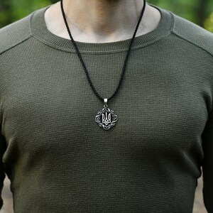 May include: A man wearing a green long-sleeved shirt with a black cord necklace and a silver pendant featuring the Ukrainian trident symbol.