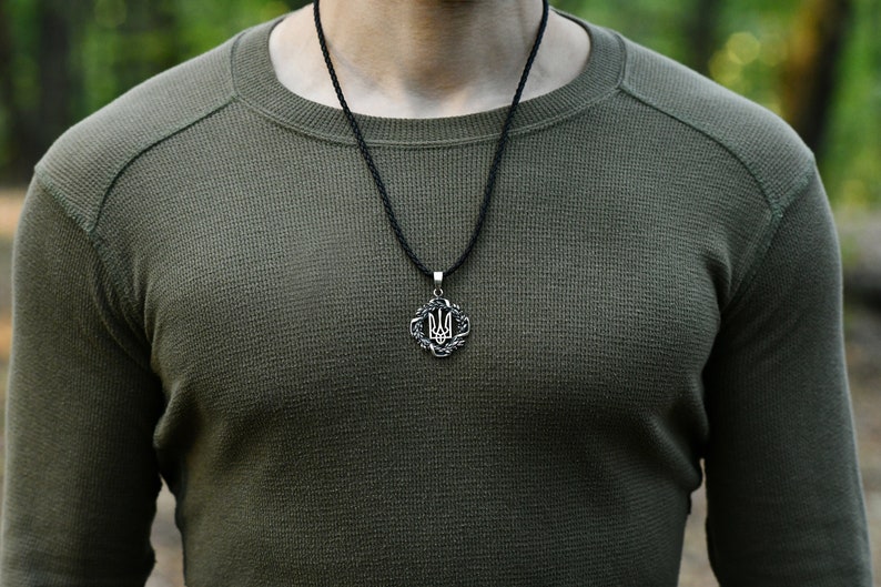 May include: A man wearing a green long-sleeved shirt with a black cord necklace and a silver pendant featuring the Ukrainian trident symbol.
