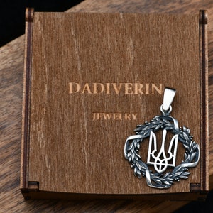 May include: A silver pendant featuring the Ukrainian trident symbol surrounded by a wreath of leaves. The pendant is in a wooden box with the inscription "Dadiverin Jewelry".