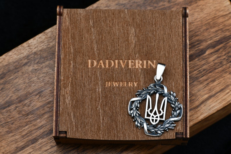May include: A silver pendant featuring the Ukrainian trident symbol surrounded by a wreath of leaves. The pendant is in a wooden box with the inscription "Dadiverin Jewelry".