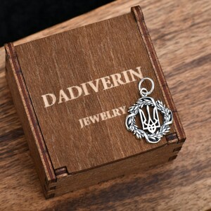 May include: A silver pendant in the shape of a wreath with a trident inside. The pendant is sitting on a wooden box with the words "Dadiverin Jewelry" engraved on the top.