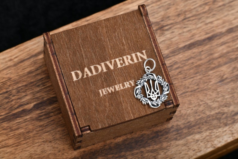 May include: A silver pendant in the shape of a wreath with a trident inside. The pendant is sitting on a wooden box with the words "Dadiverin Jewelry" engraved on the top.