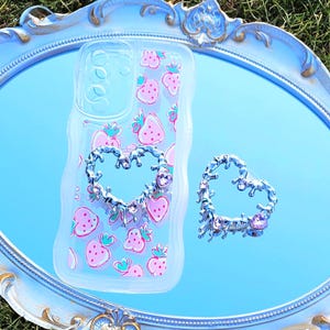 May include: A clear phone case with a pink strawberry pattern and a silver heart-shaped phone grip sits on a light blue oval mirror. A matching silver heart-shaped ring is next to the phone case. The mirror is on a light blue decorative tray with gold accents.