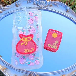May include: A clear phone case with a strawberry pattern and a red money bag design. Next to it is a red case with gold coins. The items are on a silver ornate tray.
