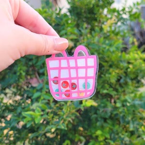 May include: A pink, grid-patterned tote bag-shaped acrylic charm. The charm is filled with colourful, circular designs, including red tomatoes and blue buttons. The charm is held up against a backdrop of green foliage.