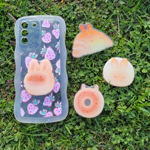 May include: A clear phone case with a pink strawberry pattern and a 3D bunny. Beside it are four food-themed stickers: a croissant, a donut, and two bunny-shaped designs. All items are on a bed of green grass.