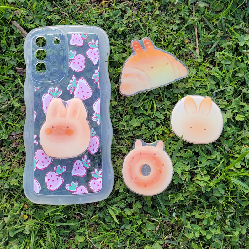 May include: A clear phone case with a pink strawberry pattern and a 3D bunny. Beside it are four food-themed stickers: a croissant, a donut, and two bunny-shaped designs. All items are on a bed of green grass.