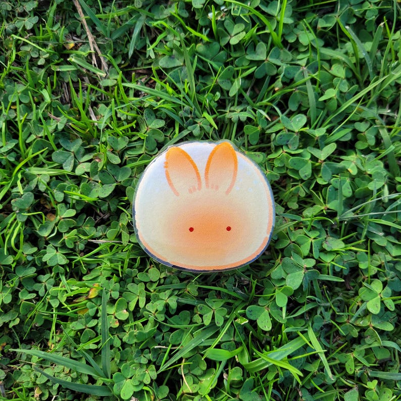 May include: A round sticker featuring a cartoon bunny face with orange ears and two small red dots for eyes. The bunny is set against a gradient of white to orange. The sticker is on a bed of green grass and clover.