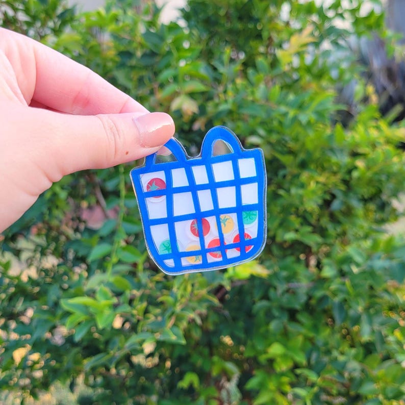 May include: A blue and white acrylic charm shaped like a shopping basket. The basket contains colourful fruit stickers, including tomatoes and other produce. The charm has two blue handles and a grid pattern.