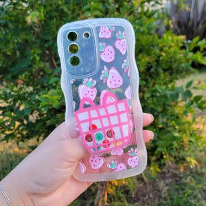 May include: Clear phone case featuring a pink strawberry pattern and a pink shopping basket design. The case has a clear border and is held in a hand against a green backdrop.